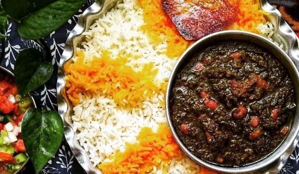 A bowl of dark green Ghormeh Sabzi served with saffron basmati rice and a wedge of lemon.